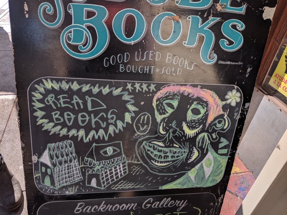 A chalk sign from Adobe Books showing a grinning monkey surrounded by stars and flowers, looking at two houses, one of which has an eye and teeth, and it says READ BOOKS