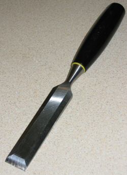 A chisel, which has a rounded wood handle about 3" long out of which a large piece of metal about 5" long protrudes. The piece of metal has a square end with an obvious bevel where its cutting edge is.