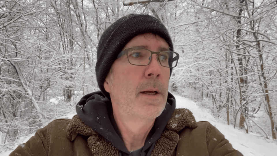A man with glasses in a knit hat walks down a snowy path, surrounded by trees covered with snow.