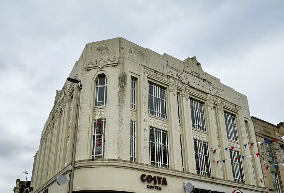 A three storey shop on a corner plot, with the photo focusing on the top two floors. The windows run the height of both floors, with the floor plate hidden by stonework. There is a lot of detail which will be described in the close-up photos that follow.