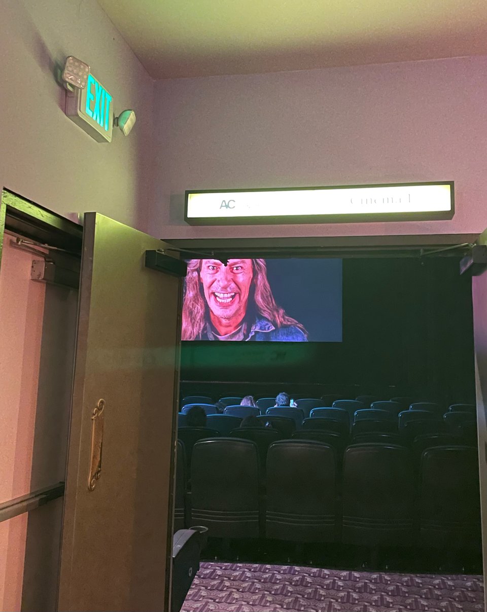A theater screening room from outside. Inside the theater, a terrifying picture of the villain Killer Bob is on the screen before the show. Outside the door, the green exit sign glows