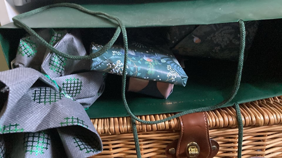 A green bag lies horizontally on to of a wicker basket. The bag is filled with presents ready to gift.