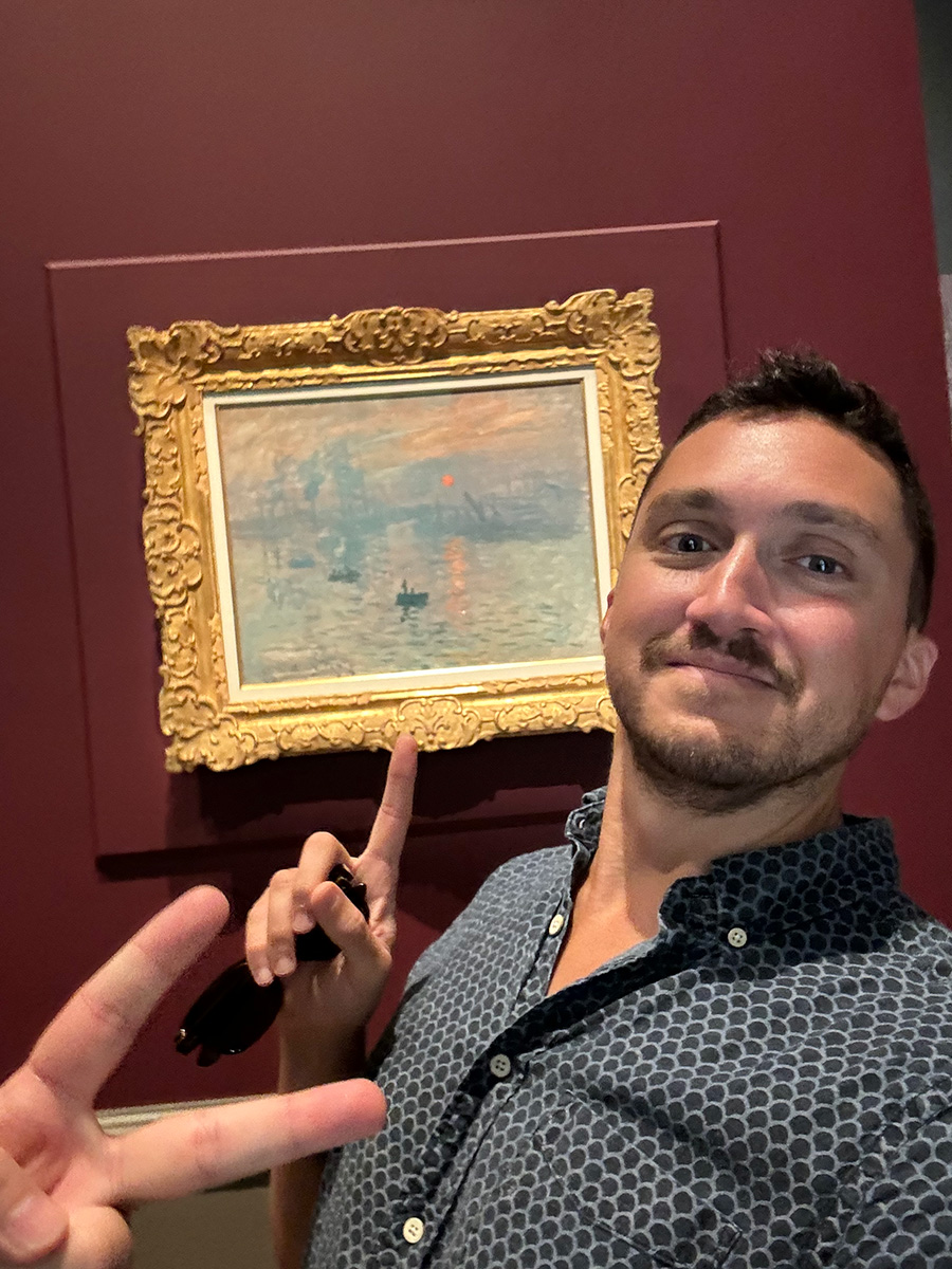 One hand in the peace sign in front of the author posing in front of a Monet painting at an art gallery.