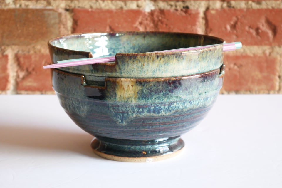 Two large ceramic bowls neatly nested, with chop sticks set in shallow notches on the rim of the top bowl. The bowls have a deep foot ring that angles one at down, then back in for the bottom quarter inch which is left bare in a speckled tan clay.