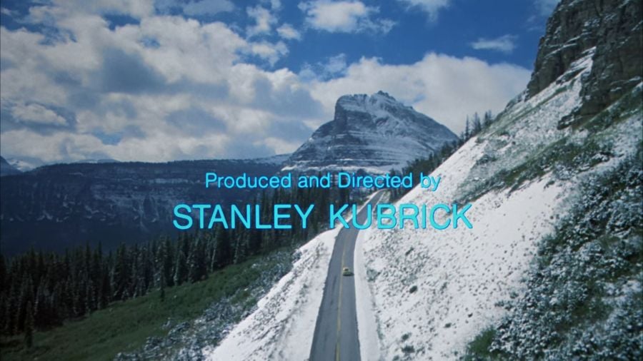 A screenshot from The Shining, showing Jack Torrance’s car winding through the Colorado mountains, just starting to reach a high enough altitude for snow to be gathering on one side of the road. There’s a snow-capped peak in the background, and text overlaid reading “Produced and Directed by Stanley Kubrick”