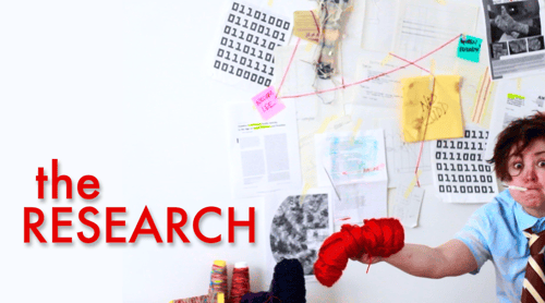 a disheveled person, dressed in a short-sleeved shirt and tie, holding a seam-ripper between their lips like a cigarette, stands in front of a conspiracy wall covered in documents related to knitting machines, sticky notes, a stained napkin with 'NED LUDD' scrawled across it, and criss-crossed red string; they wave a skein of bright red yarn towards the camera while making intense eye contact; a red text overlay reads 'the research'