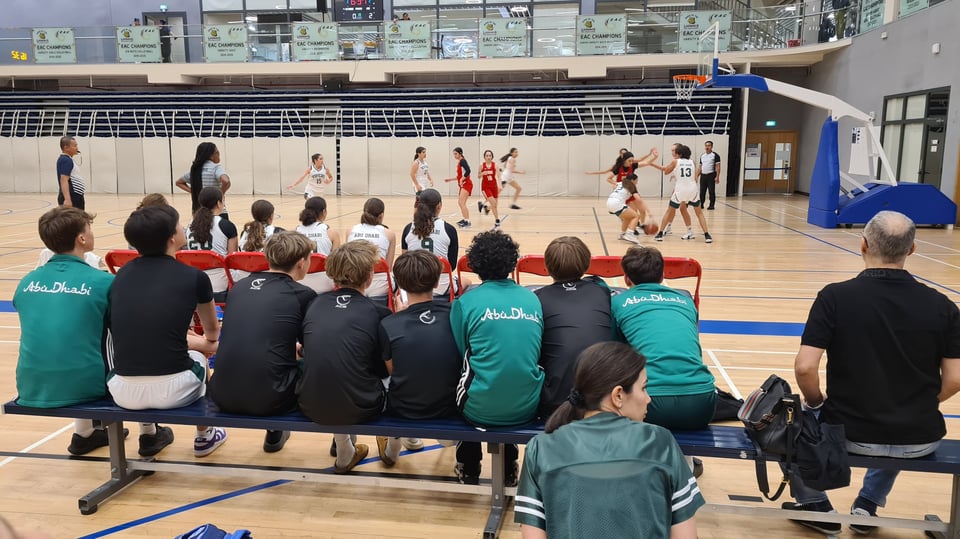 The JV boys basketball team supportinf the girls' team. They are seated in a row along a bench as the game goes on.