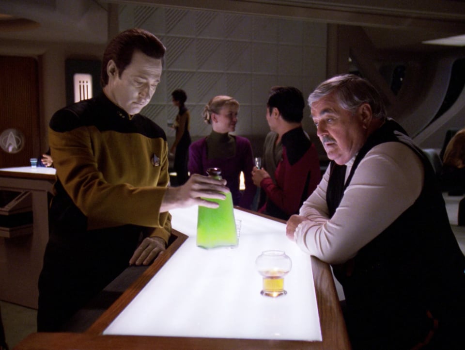 Data and Scotty are at the bar at Ten Forward. Data is holding a glass bottle of bright green liquid. Scotty eyes the bottle, hopefully.
