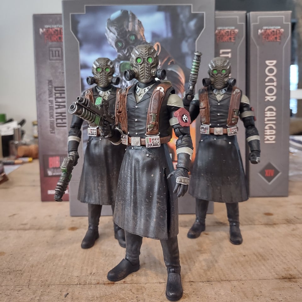 Three action figures stand in loose formation on a wooden table. They all wear long coats, each with a heavy leather harness over it. Their heads are completely covered by retro-futuristic gasmasks with large green eye lenses.