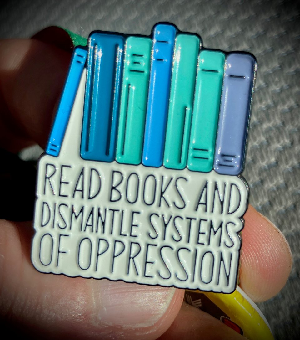 Fingers hold a new pin. At the top a few books as they sit on a shelf. Under the books a text that reads: Read books and dismantle systems of oppression.