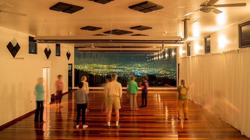 A group of people getting ready for a qigong class, in a room with a wood floor and big picture windows showing a view of San José's city lights in the distance