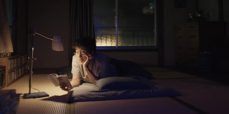 An elderly Japanese man reads a book in his apartment on a low bed.