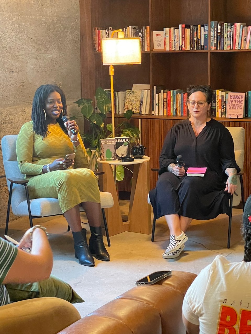 Hannah Eko and Wendy C. Ortiz are seated in chairs. Bookshelves are behind them. In between them is a small table with a lamp overhead, and the books Hollywood Notebook and Bruja are facing out.