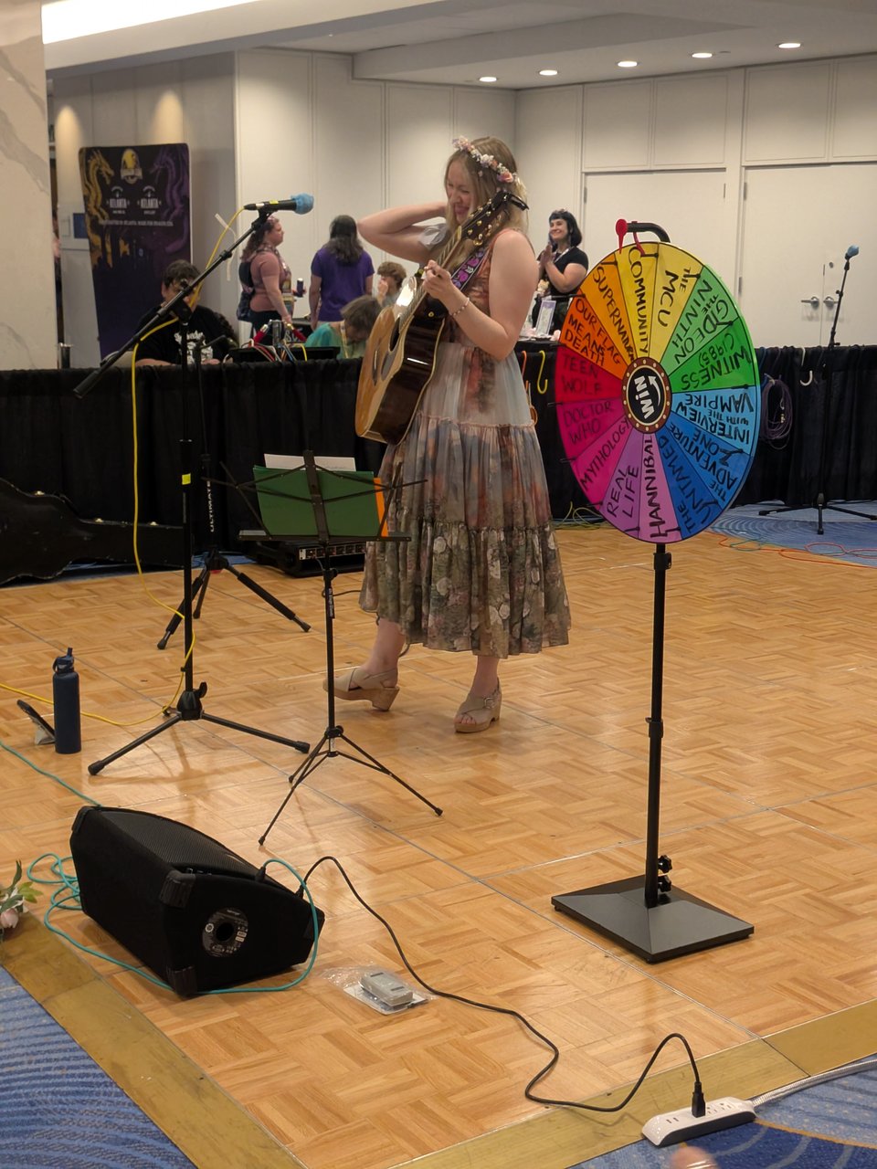 A photograph of Emily Henry on the Concourse Stage at Dragon Con, holding a guitar and standing next to a multicolor prize wheel.