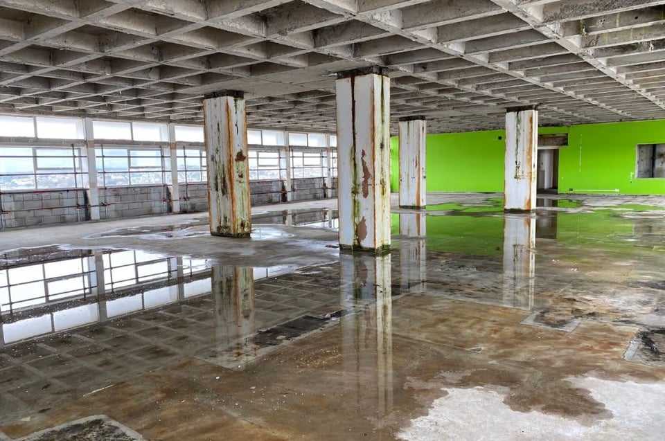 An empty floor of the civic centre. The suspended ceilings and flooring has been stripped out to reveal the coffered ceiling and bare concrete floor. Several square columns show signs of peeling paint and rusted edges and the floor has pools of water on it.