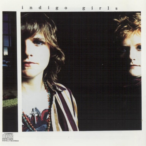 A color photgraph of two white women, Indigo Girls, Amy Ray on the left and Emily Saliers on the right. Ray has shoulder length brown hair and is wearing a crucifix; half her face is cropped out of the shot. Saliers has short blond hair, is lit from the right side, and but otherwise is in darkness.
