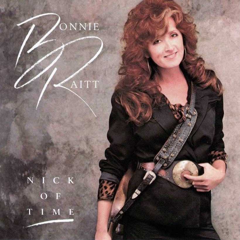 A photograph of Bonnie Raitt, a lean white woman with big curly reddish brown hair with a slight streak of white on the front. She's dressed in black, possibly leather, and the shirt underneath has a tiger pattern print. She's smiling. Slung around her shoulder is her guitar, but the guitar is on her hip and mostly cut out of the picture, so we only really see the strap, which looks vaguely like snake skin. One hand is on the guitar, and the other is on her giant metal circular belt buckle. The belt buckle has a red-brown circle in the middle, and she has her finger on it, like pressing a button.