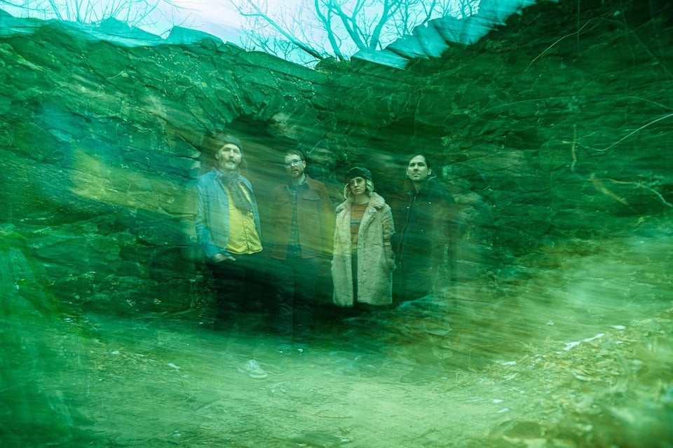 Four people in winter clothing stand in front of what resembles a ruined stone structure. The photo has a hazy overlay, giving it a greenish tint that evokes the music on the band's album.
