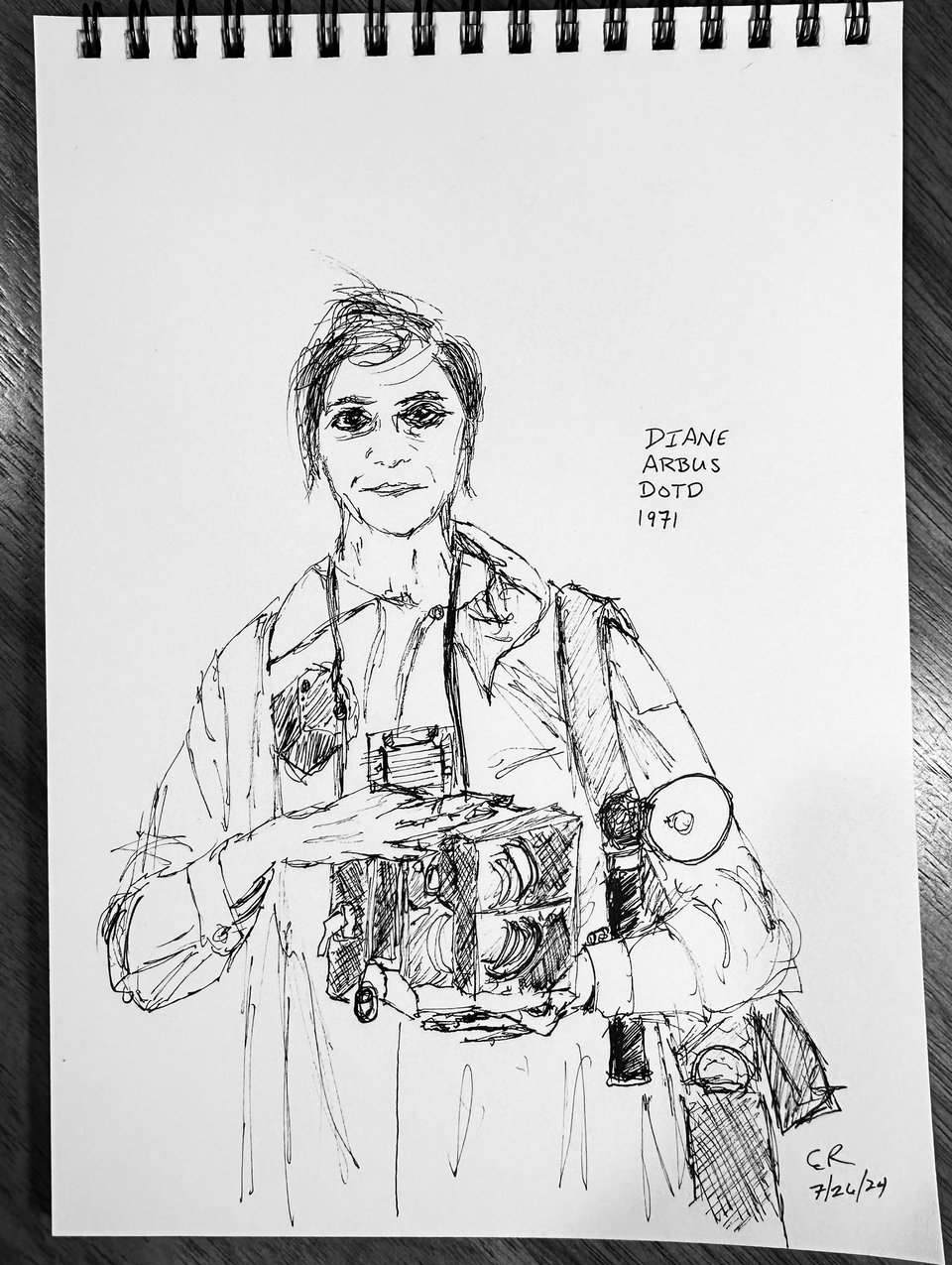Photo of sketchpad drawing of photographer Diane Arbus, in a raincoat holding photographic equipment