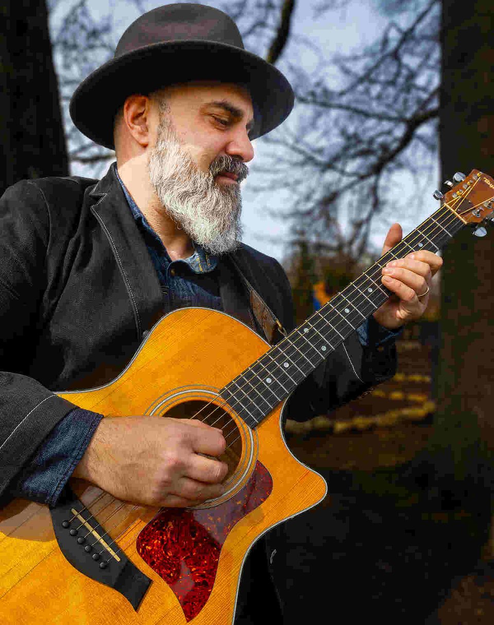 A man with a graying beard wearing a fedora plays an acoustic guitar, flanked by trees with bare branches.
