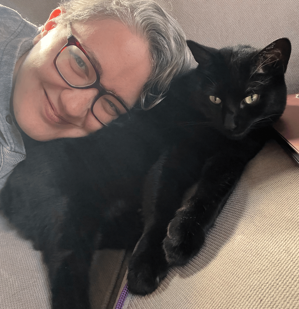 Becky, with silver hair and glasses, resting their head on the side of a very tolerant black cat. The cat does not look impressed.
