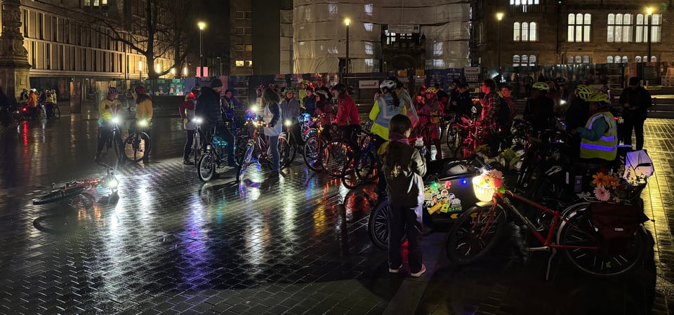 A group of around fifty cycle users - mostly women and girls - stand in the middle of Bristo Square, in a mix of brightly coloured high-visibilty clothing, bike helmets, and bikes decorated with colourful lights against the backdrop of a grey Edinburgh winter evening.