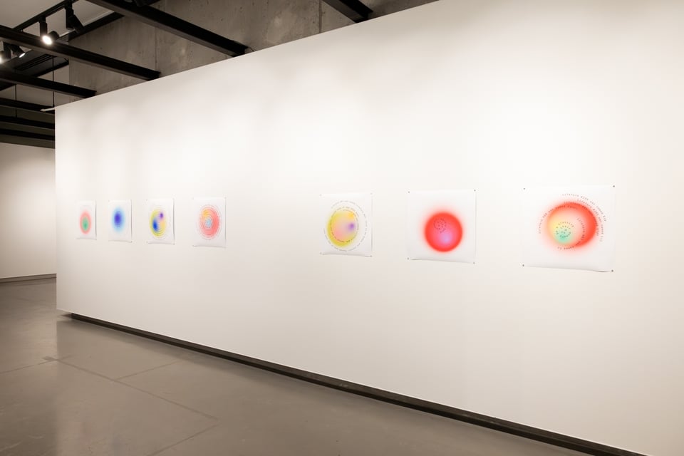 Seven printed memoryforms hung in a row on a white wall in a brightly lit gallery. They are soft orbs in pastel colours, some simply (just one colour) and others with many smaller circles of colour inside, like a petri dish