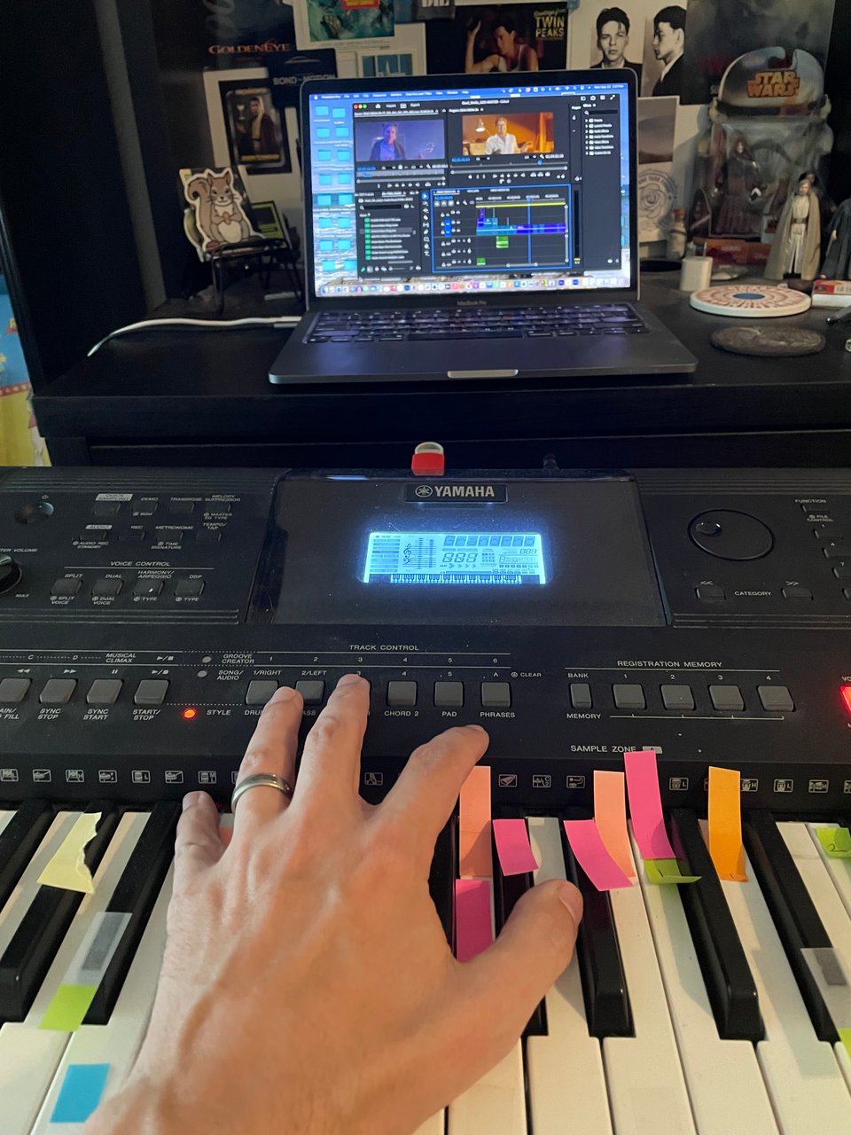 A cluttered desk, a laptop with editing software on it. A keyboard with various keys covered in color coded post-its and a hand reaching out to play them