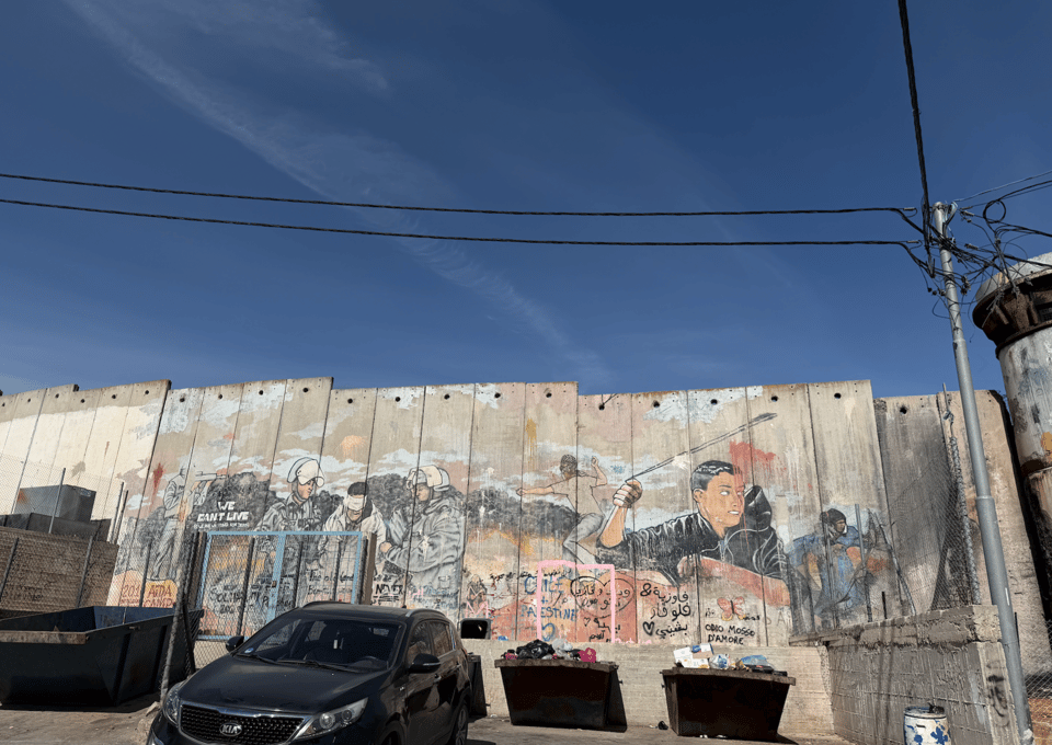Photograph of a tall concrete barrier wall featuring a large mural depicting a confrontation between a young man and soldiers, with the man holding a sling shot and police in helmets and shields. The mural includes various colors and Arabic text, and the words in English WE CAN’T LIVE. In front of the wall is a black car and trash bins  under a clear blue sky, with electrial wires above the wall. A tall watchtower is seen at the edge of the image