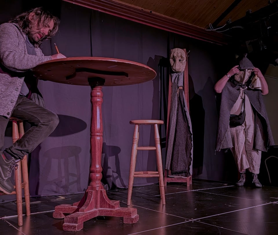 A dimly lit theatre or rehearsal space bathed in pink-purple stage light, where a long-haired man in a grey cardigan sits on a wooden stool writing with a pencil at a round pedestal table, an empty stool stands beside him, and at the right a performer in layered grey and beige robes adjusts a mask over their face beside a costume stand holding another mask and draped fabric, with a black-curtained backdrop and taped grid markings on the dark floor.