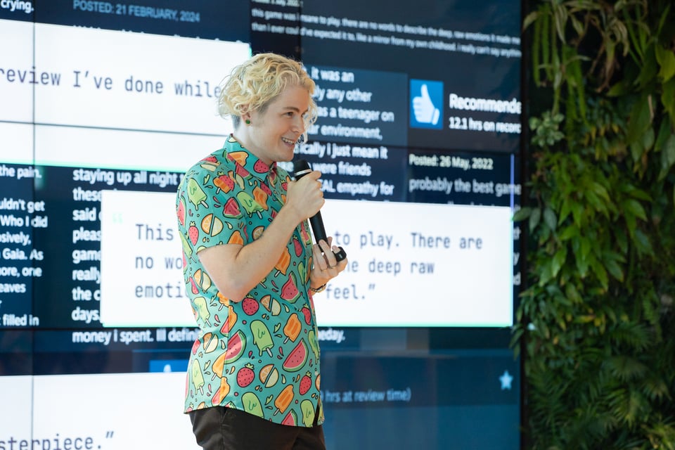 A blonde, white person giving a talk on stage. They are smiling and standing in front of a large screen.