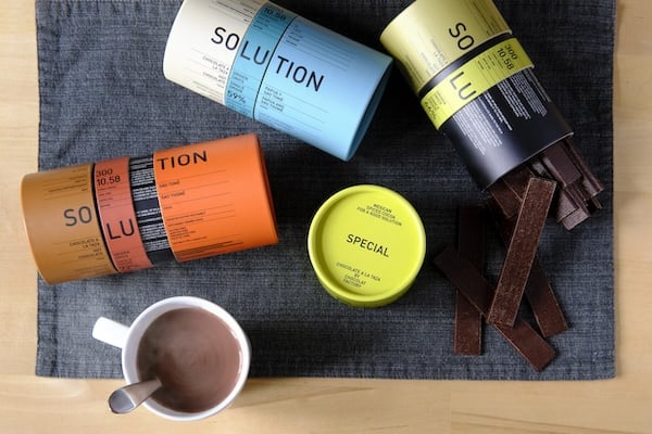 Sobre un mantel de color gris oscuro, una taza de cocholate y tres packs de Solution. Son cilíndricos y se separan en tres partes de manera que la palabra Solution queda separada en tres sílabas. Uno de los packs es naranja y marrón, otro azul y blanco y un tercero es verde y negro.