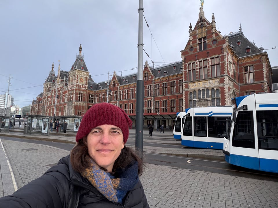 A woman wearing a red knit cap, thick blue scarf and a puffy black jacket stands in front of the Amsterdam Central Train station. Three trams stand in the background