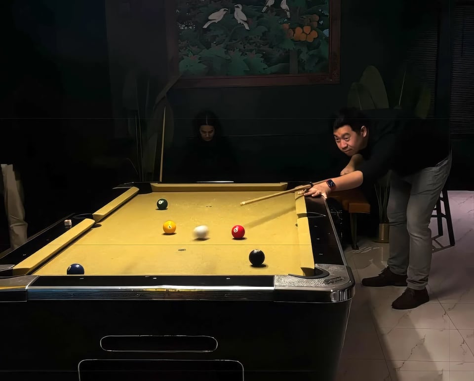 A man in a black shirt and gray pants leans over a mustard-colored pool table, aiming his cue for a precise shot. The white cue ball is in motion toward several colored balls scattered across the table. In the dimly lit background, a woman sits quietly, half in shadow, beneath a wall painting of birds and trees. The pool table is brightly lit, but everything is quite dark, making it moodily dramatic