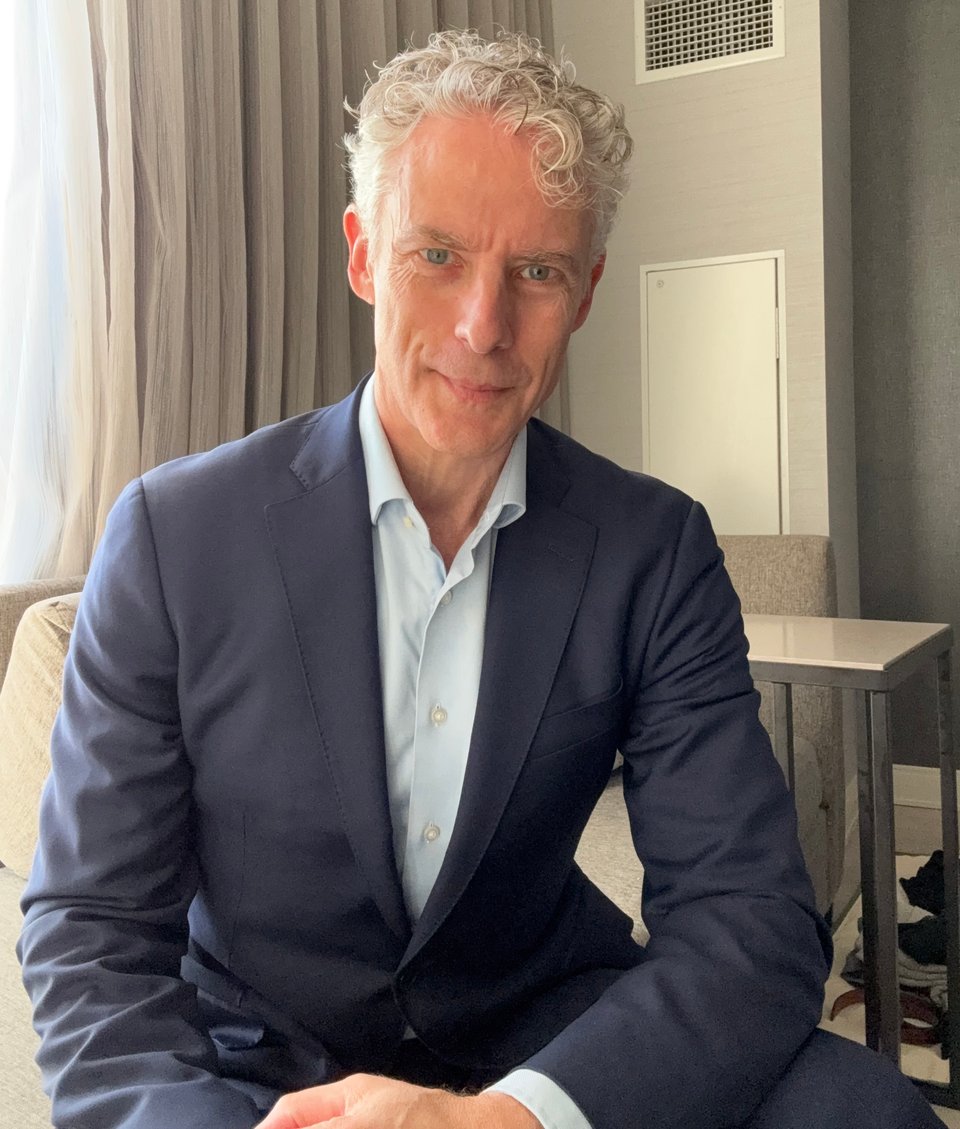 selfie of middle-aged white man in navy blue suit and light blue dress shirt with no tie, sitting in a hotel room
