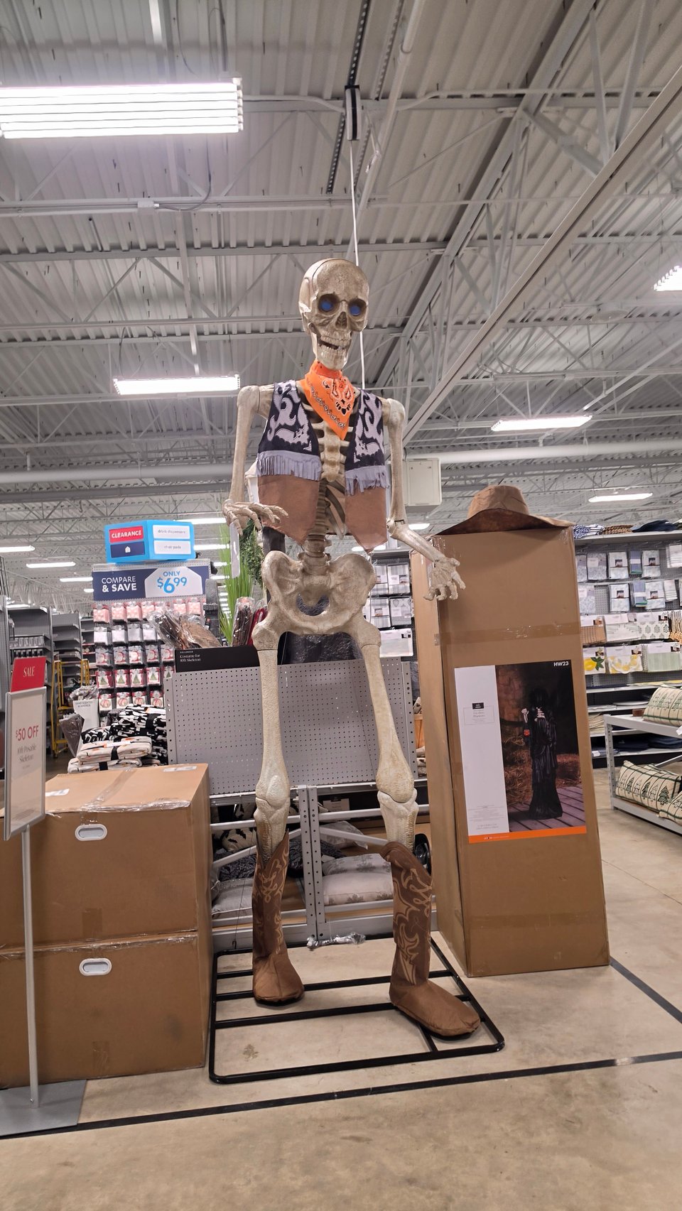 A tall skeleton Halloween decoration dressed as a cowboy with a fringed vest, orange bandana, and brown cowboy boots stands on a metal base in a store aisle, surrounded by large cardboard boxes and a clearance sign offering $50 off.