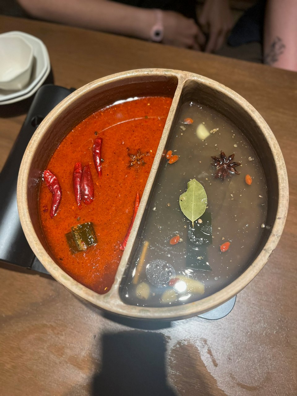 A bowl of hotpot with a divider running down the middle. On the left is a bright red meat broth, and on the right is a dark semi-opaque dashi broth. Both have kelp, star anise, and other spices floating on the surface.