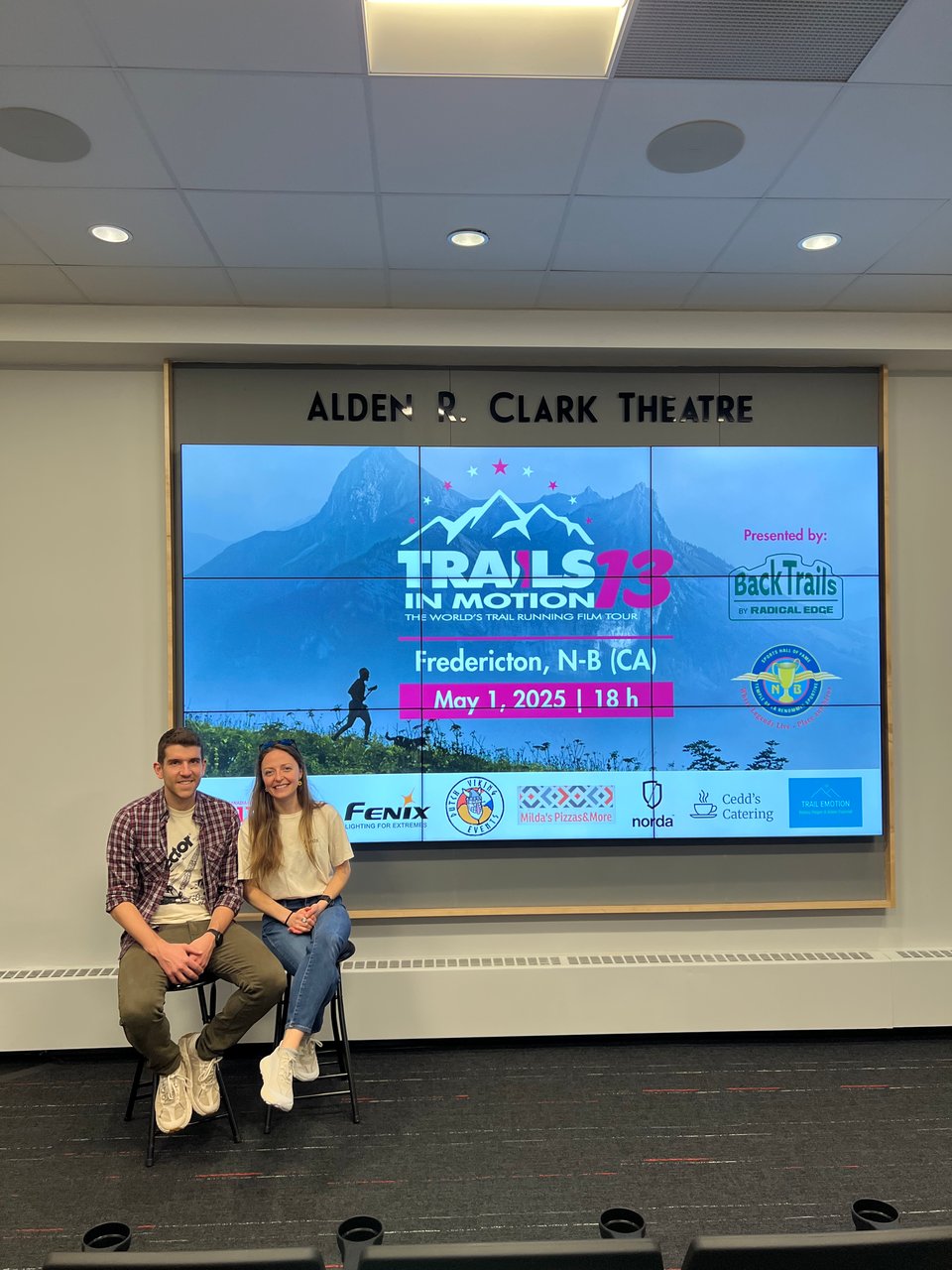 Kelsey and Adam sitting in front of a video screen at the Trails in Motion Film Festival. Adam is wearing a plaid shirt and white-ish norda shoes. Kelsey is wearing a white shirt and whiter norda shoes.