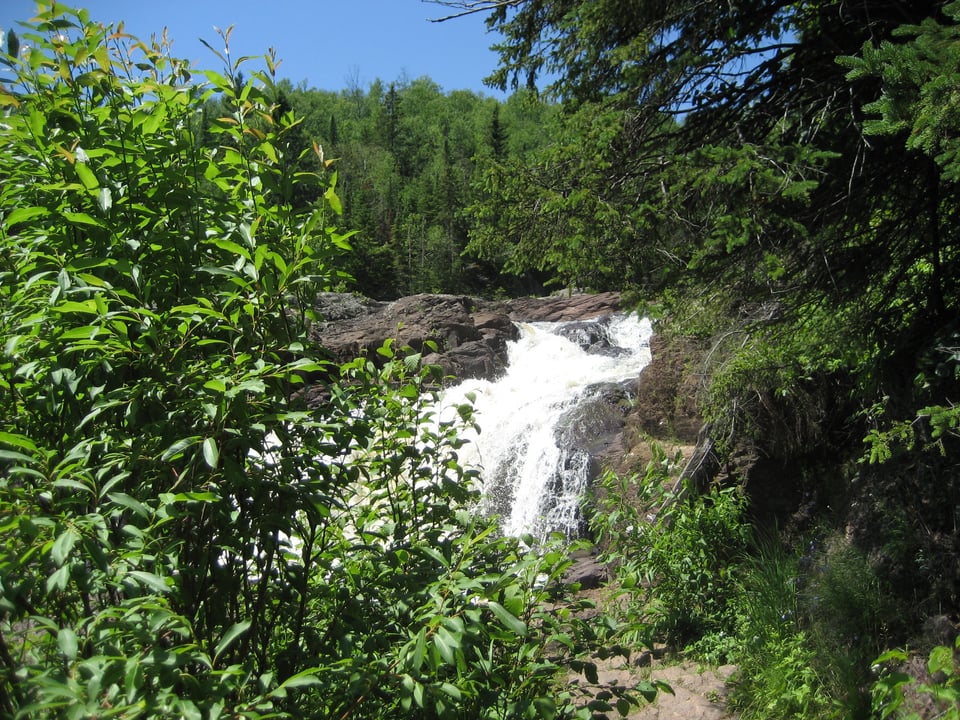 A white waterfall flows between dark grey rocks and tall pine trees.
