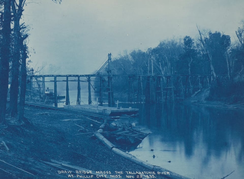 A photograph in a blue tint of a river with a rickety bridge over it and logs in the foreground.
