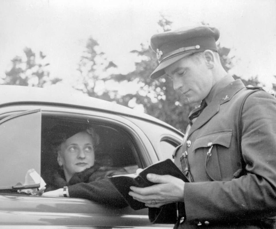 black and white photo. A man wearing a police officer uniform writes in a book. A woman in the drivers seat of a car looks on
