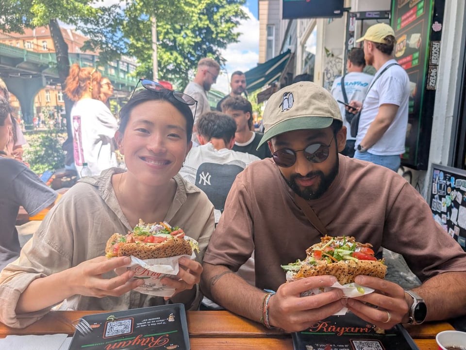 Jenny and Dillan sitting at a table holding giant burger-looking kebabs filled to the brim with vegetables and sliced meat and sauce, with a long line behind them.