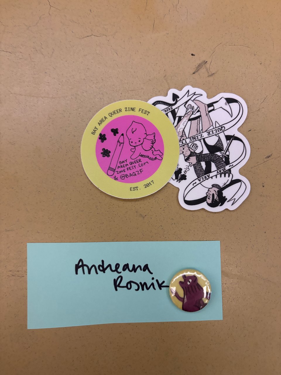 an assortment of stickers and a button pin made by Bay Area Queer Zine Fest next to the artist's name tag