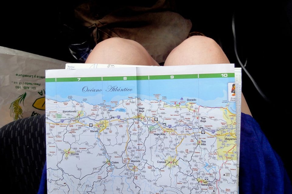 Map of coastal region on person's lap in a car, with blue ocean above and roads marked. Casual travel mood.