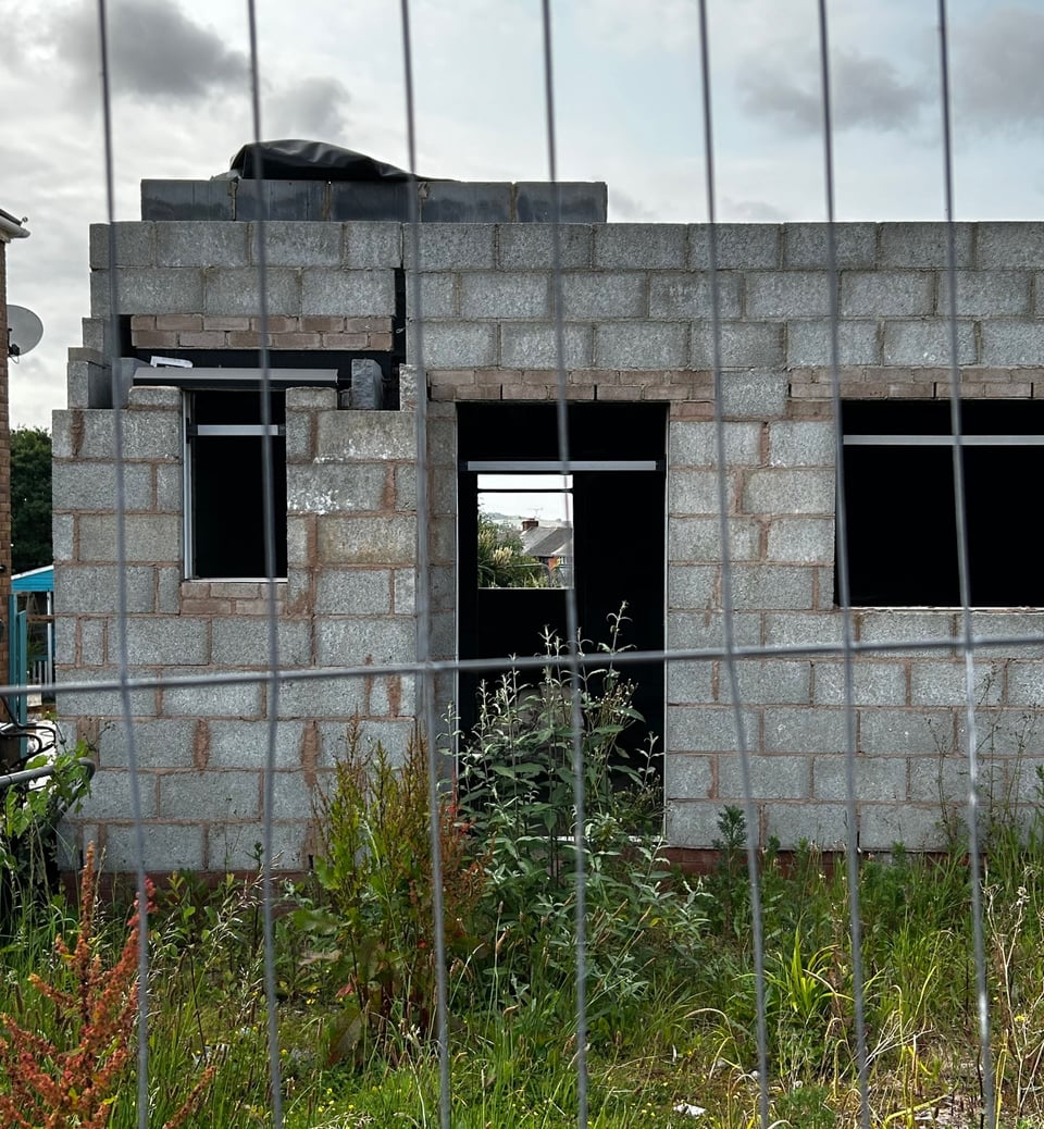 The inner shell of a the ground floor of a house surrounded by a security fence and vegetation. The walls are just concrete breezeblocks and lintels, and are braced.