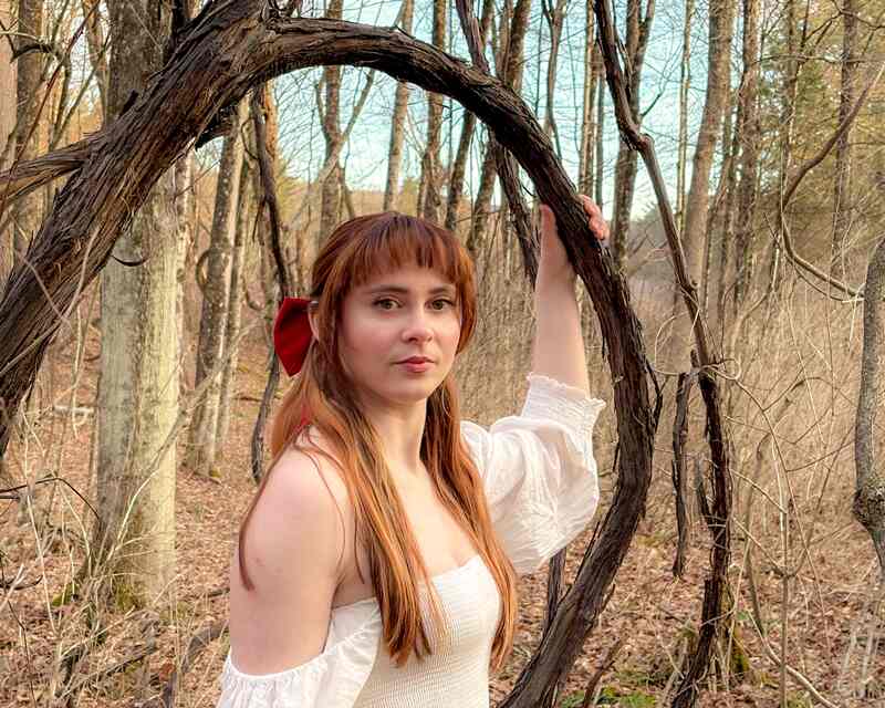 A woman from the waist up. She has long, reddish-brown hair, is wearing a white dress off her shoulders and stands in the woods with no leaves on the trees.