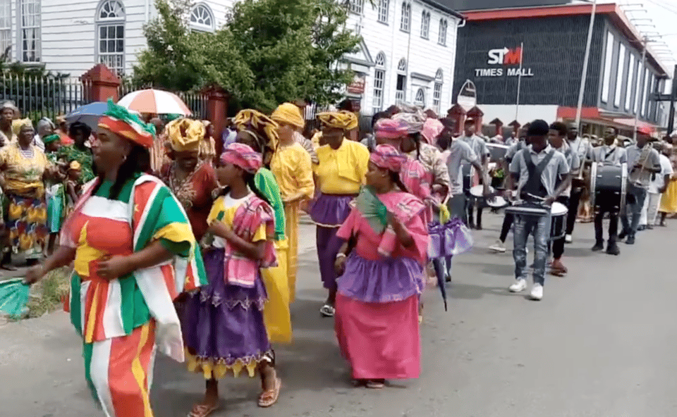 black women in bright colors processing in a street with a drum corps behind them