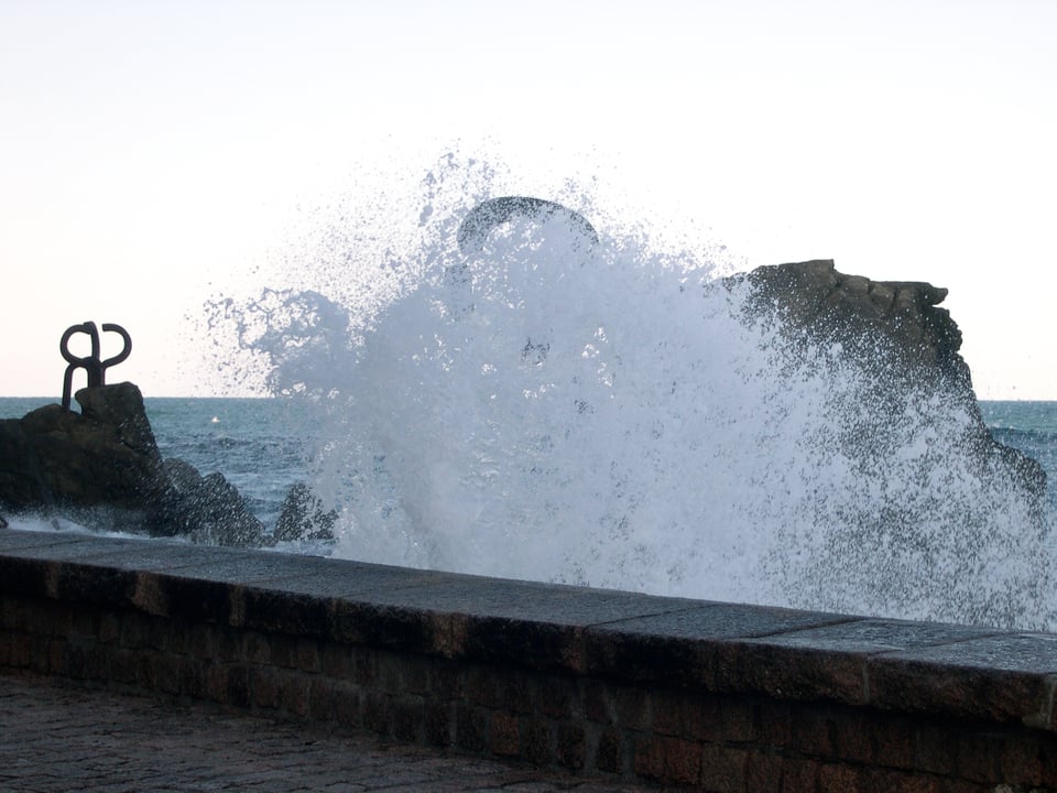 The view of the same two large steel sculptures, weighing 10 tons each, embedded in natural rocks rising from the sea, but this time a wave is breaking into the wall of the viewing area covering one of the large sculptures.