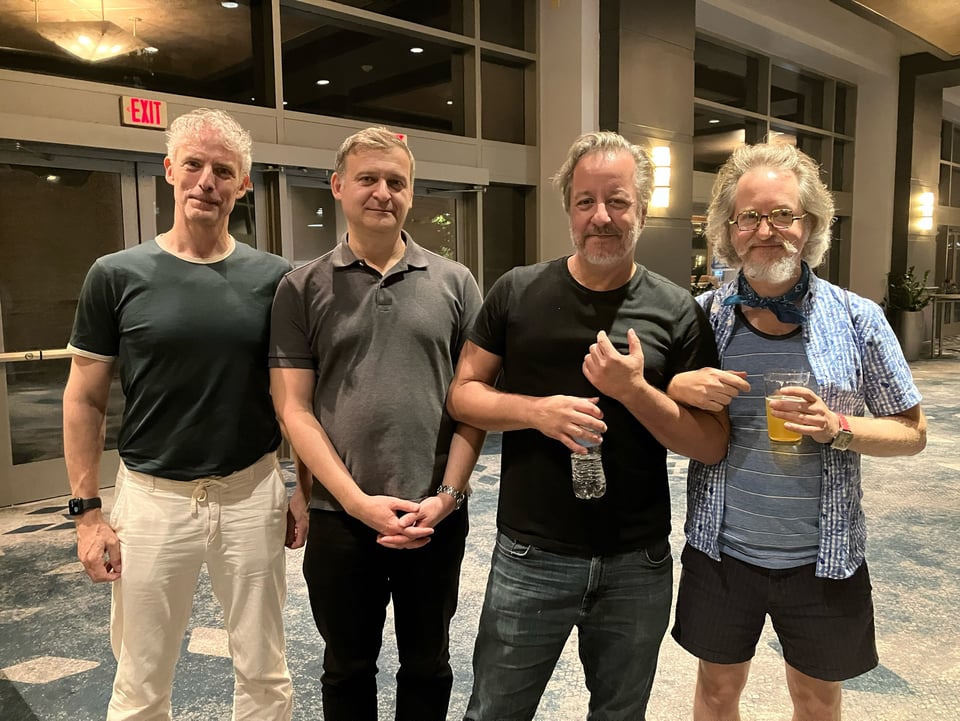 photo of 4 middle-aged white men standing side by side in a hotel conference area; 3 of them are cartoonists