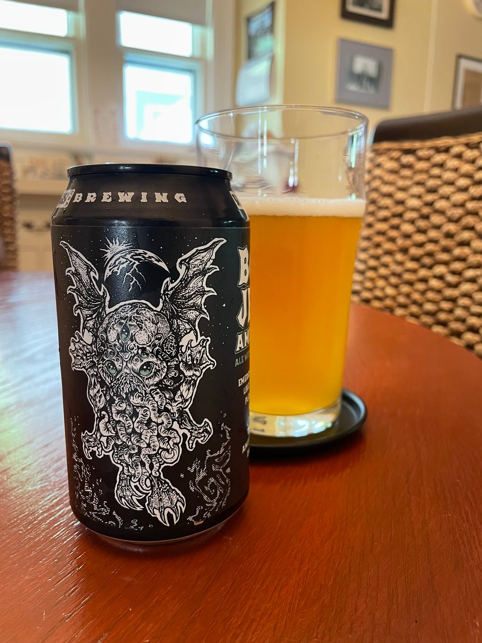 A glass of beer and a beer can sit on a wooden table on a sunny day. The beer can features a Cthulhu like monster on it